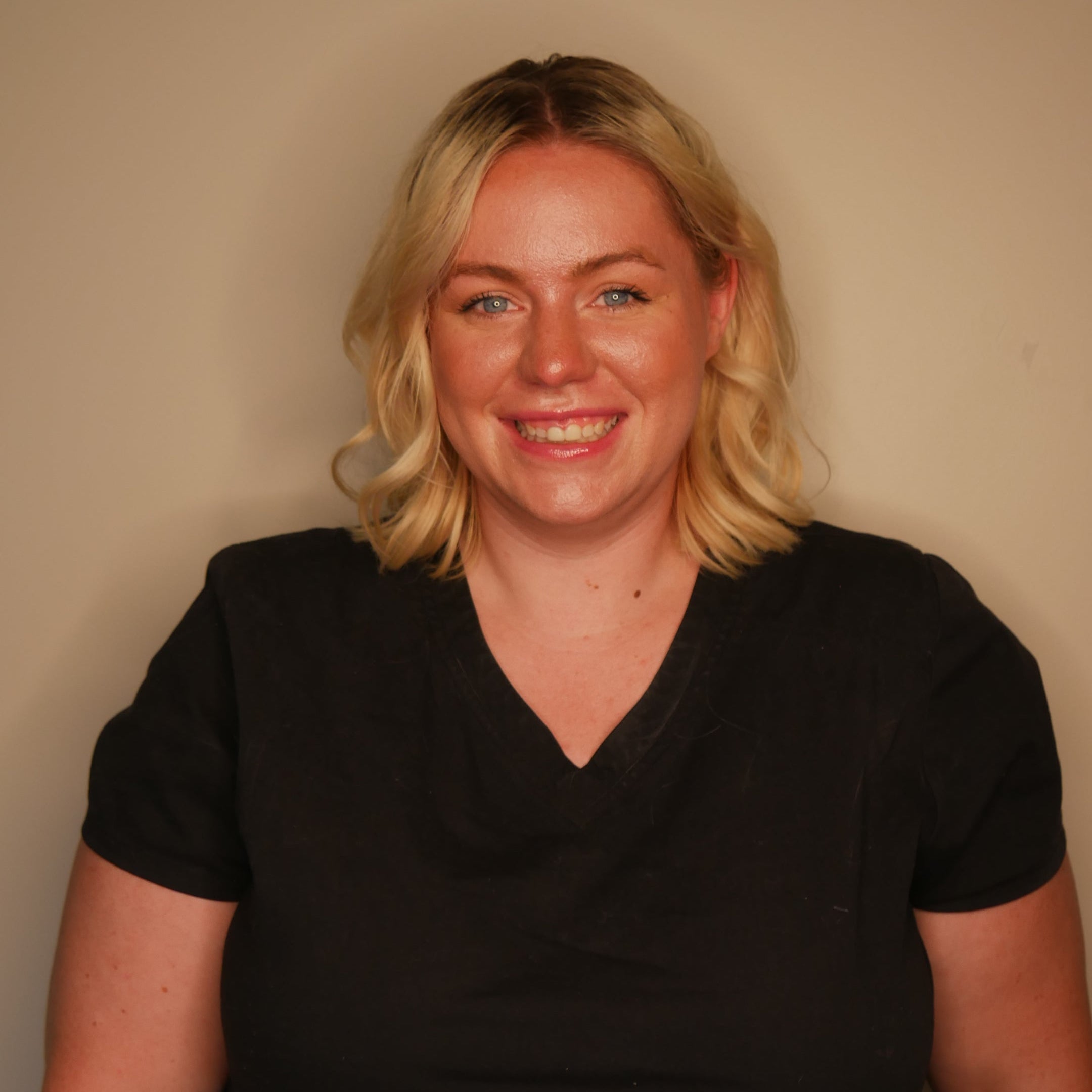 A smiling blonde woman wearing a black short-sleeve V-neck t-shirt, photographed against a plain beige wall.