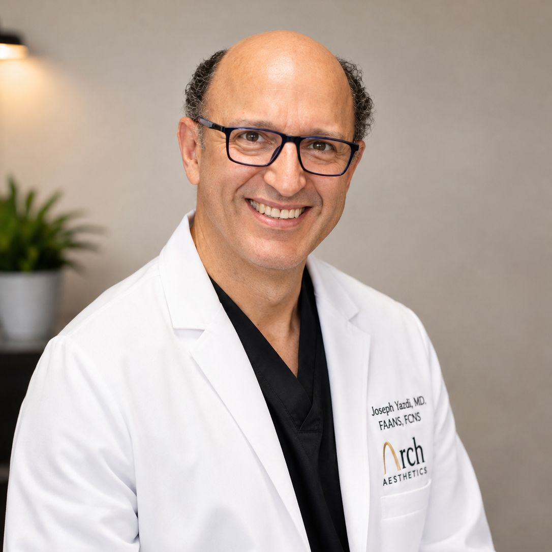 A smiling man wearing a white medical lab coat over a black scrub top, with glasses and a bald head, photographed in a professional indoor setting. The lab coat has visible text reading 'Joseph Yeshi, MD' and 'Frank Jons' with the Arch Aesthetics logo.