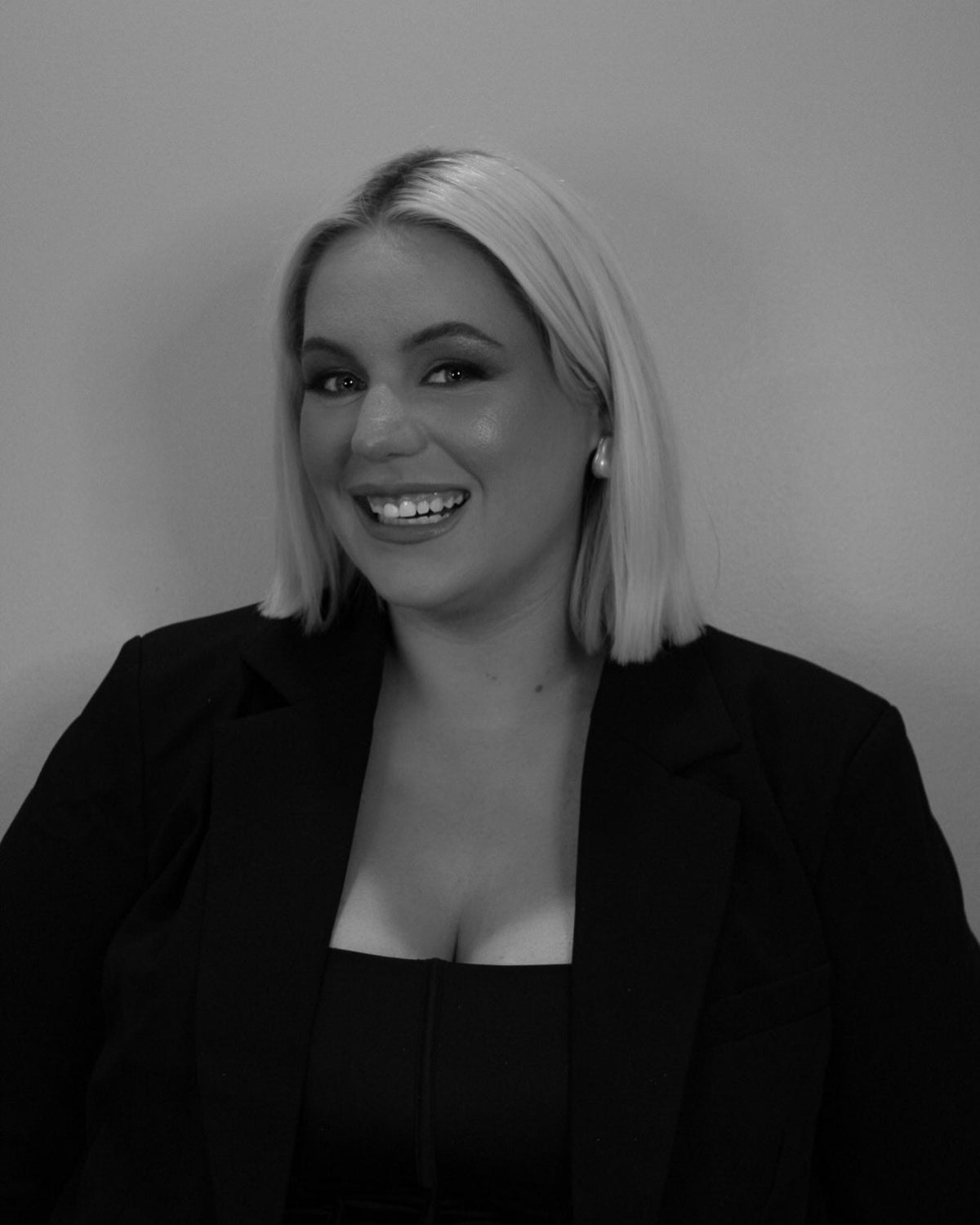 A black and white portrait of a smiling woman with shoulder-length blonde hair, wearing a black blazer over a black top against a plain light gray background.